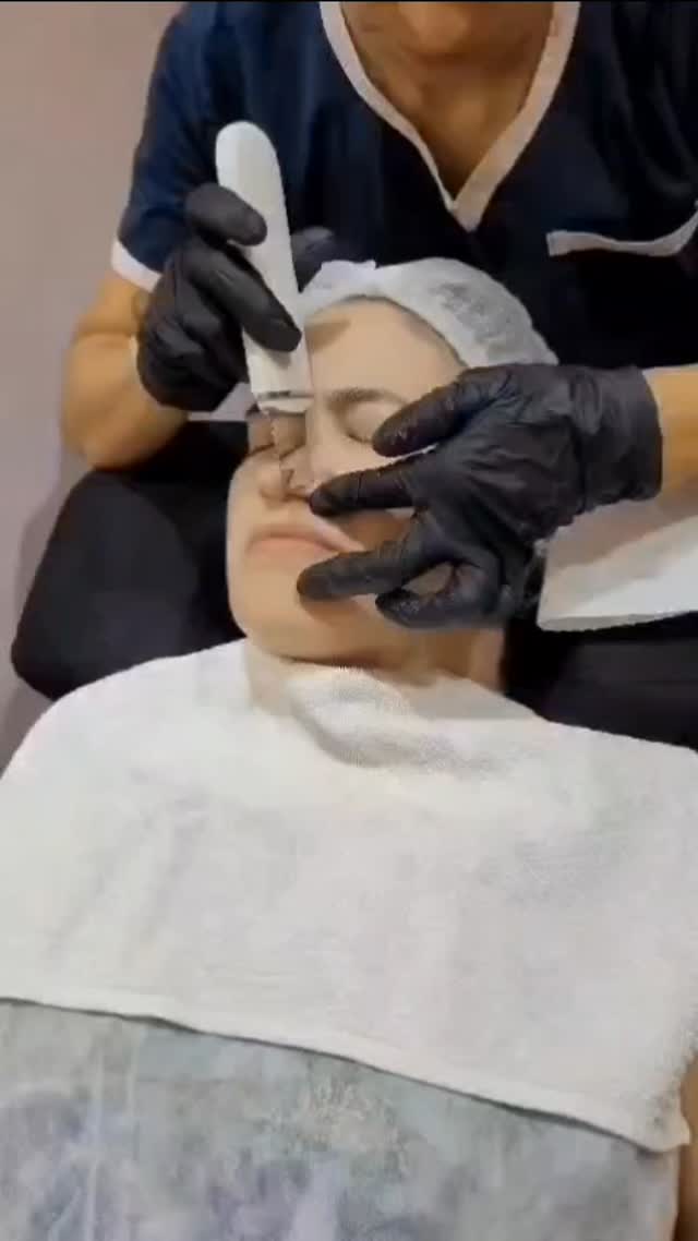 Tratamiento facial con espátula ultrasónica: manos con guantes negros sobre el rostro de una clienta en la camilla