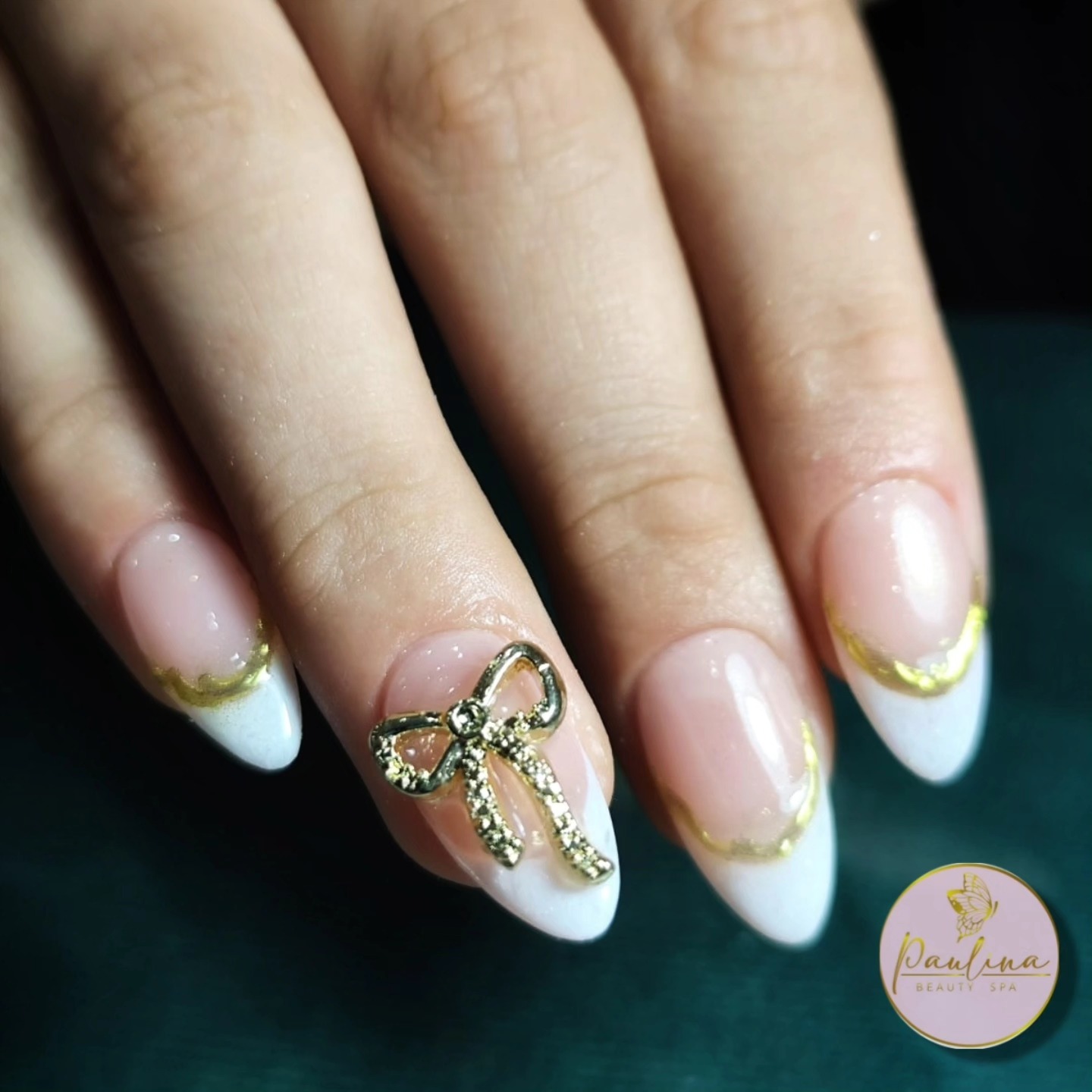 Uñas almendradas con francesita blanca, una línea dorada ondulada y un moño dorado en relieve